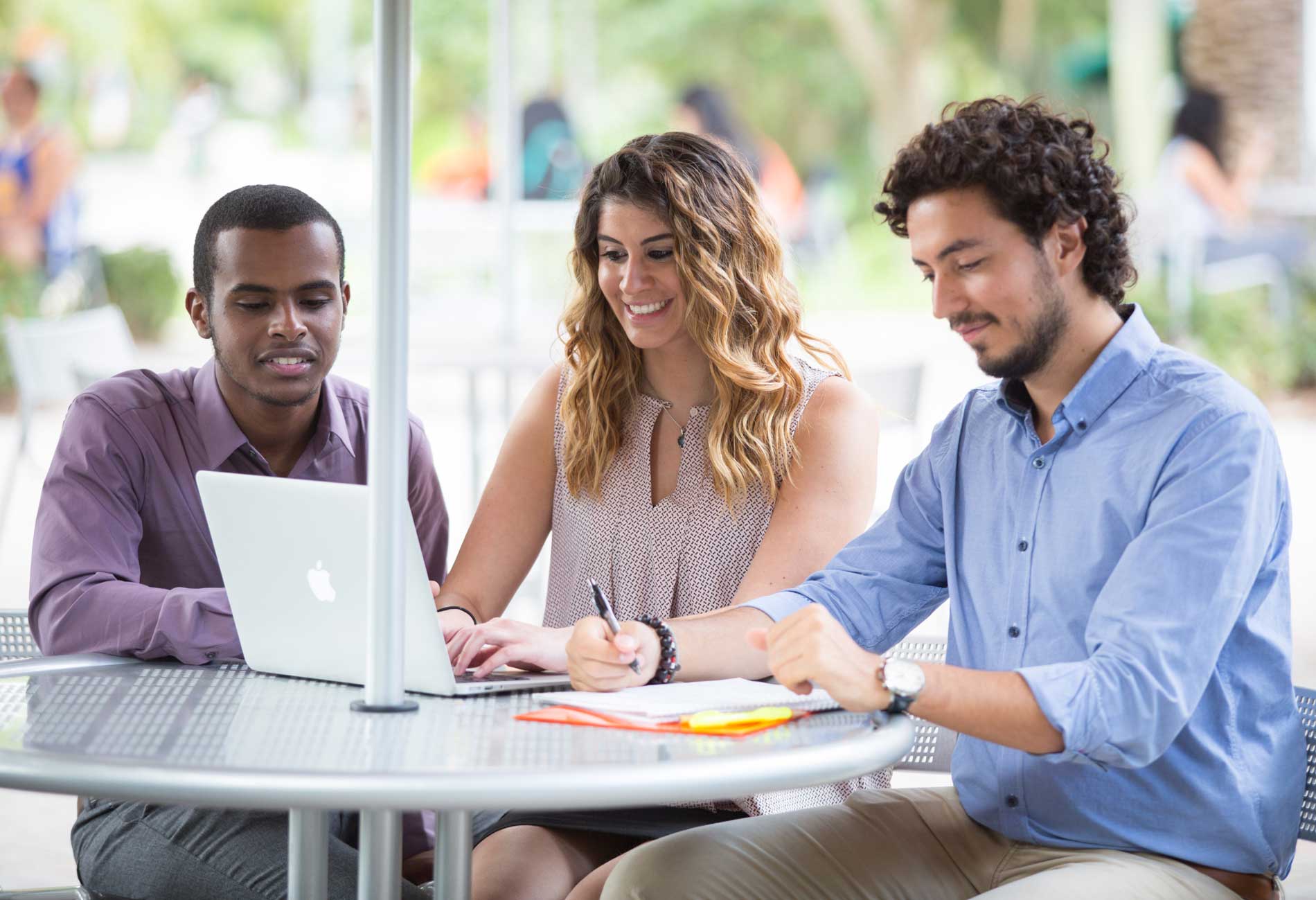 Three graduate students sitting at a table outdoors collaborating over a laptop.