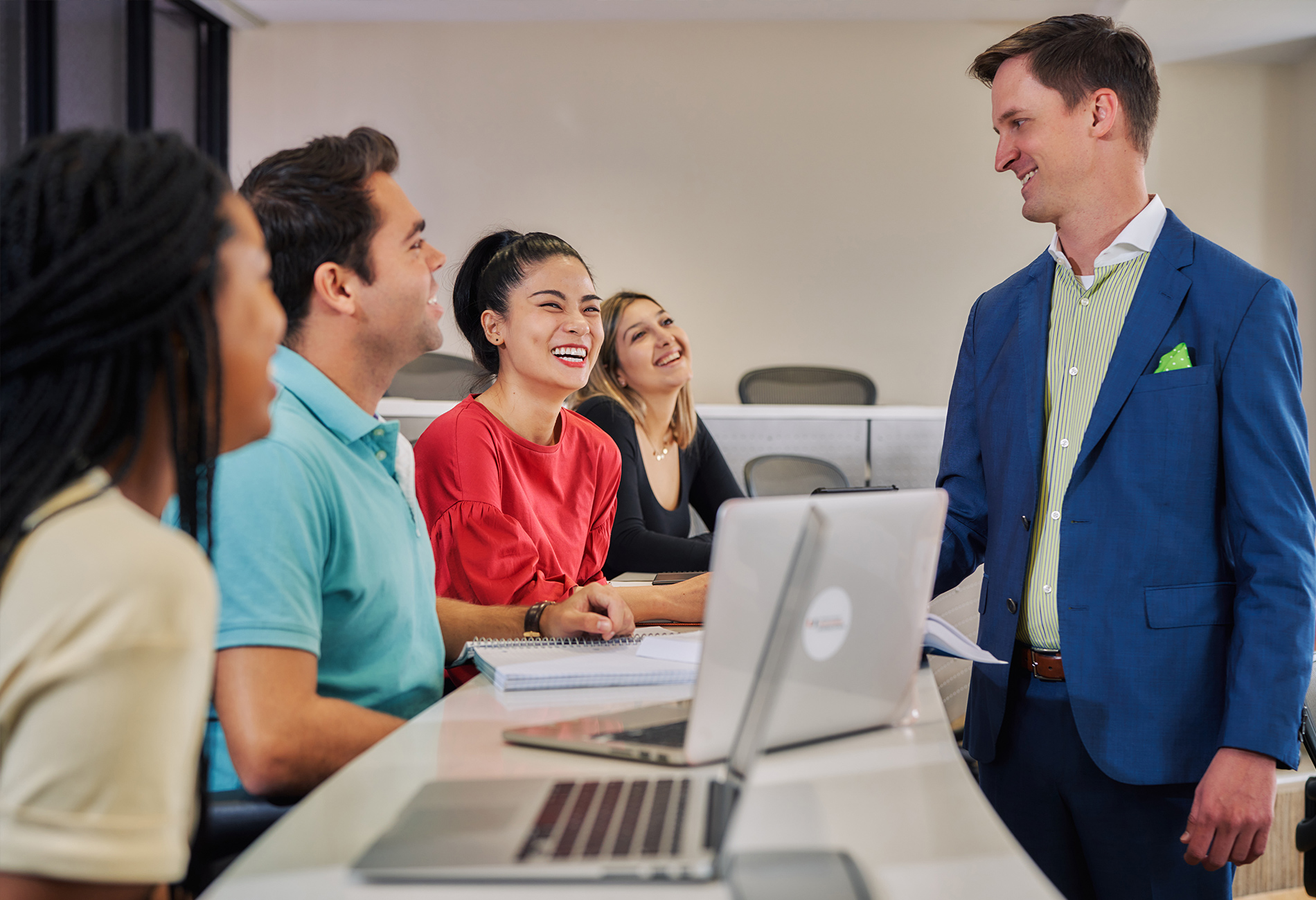 A professor interacts with four students with laptops in a classroom