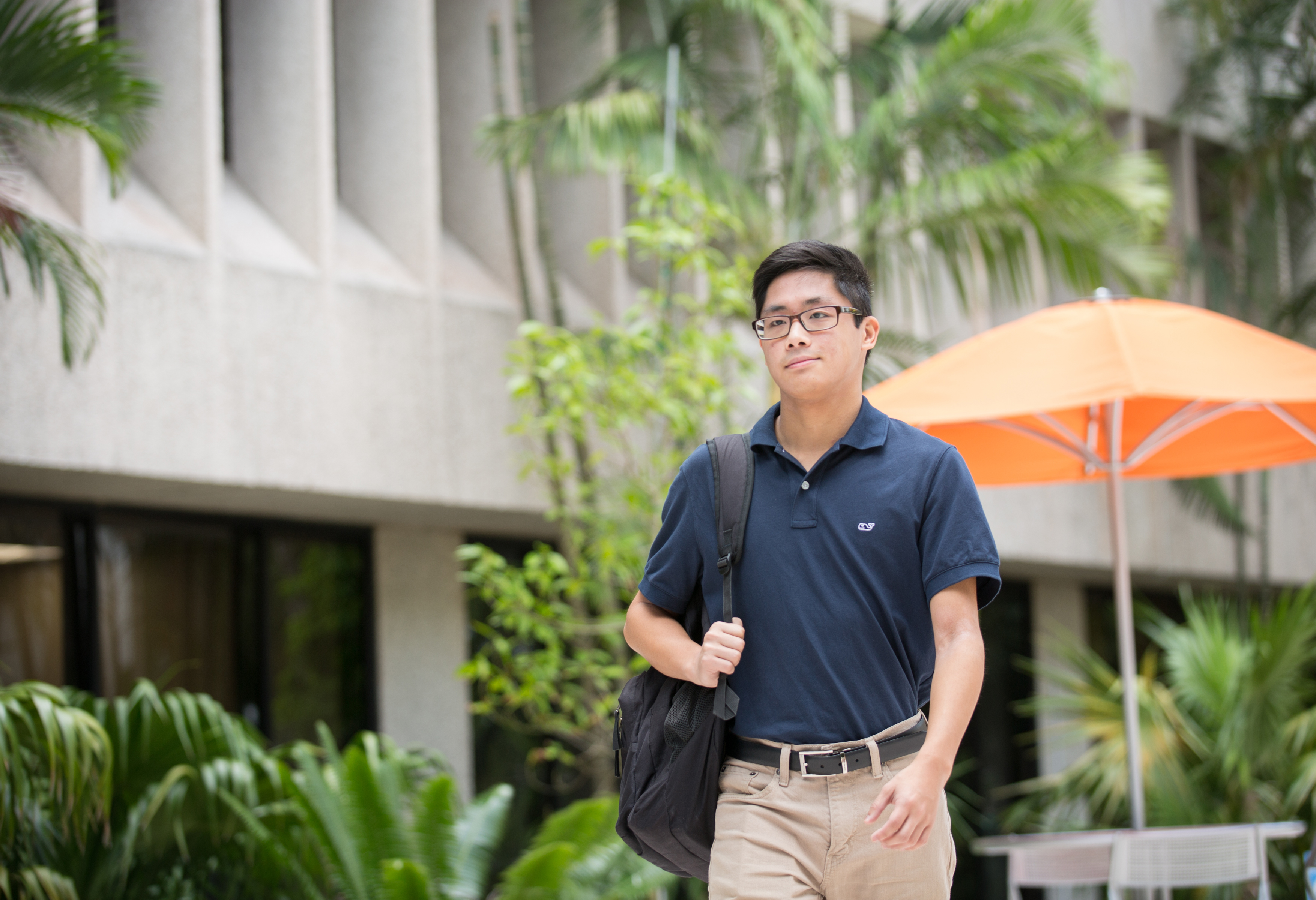 Male student walking through campus, orange umbrellas and trees in the background.