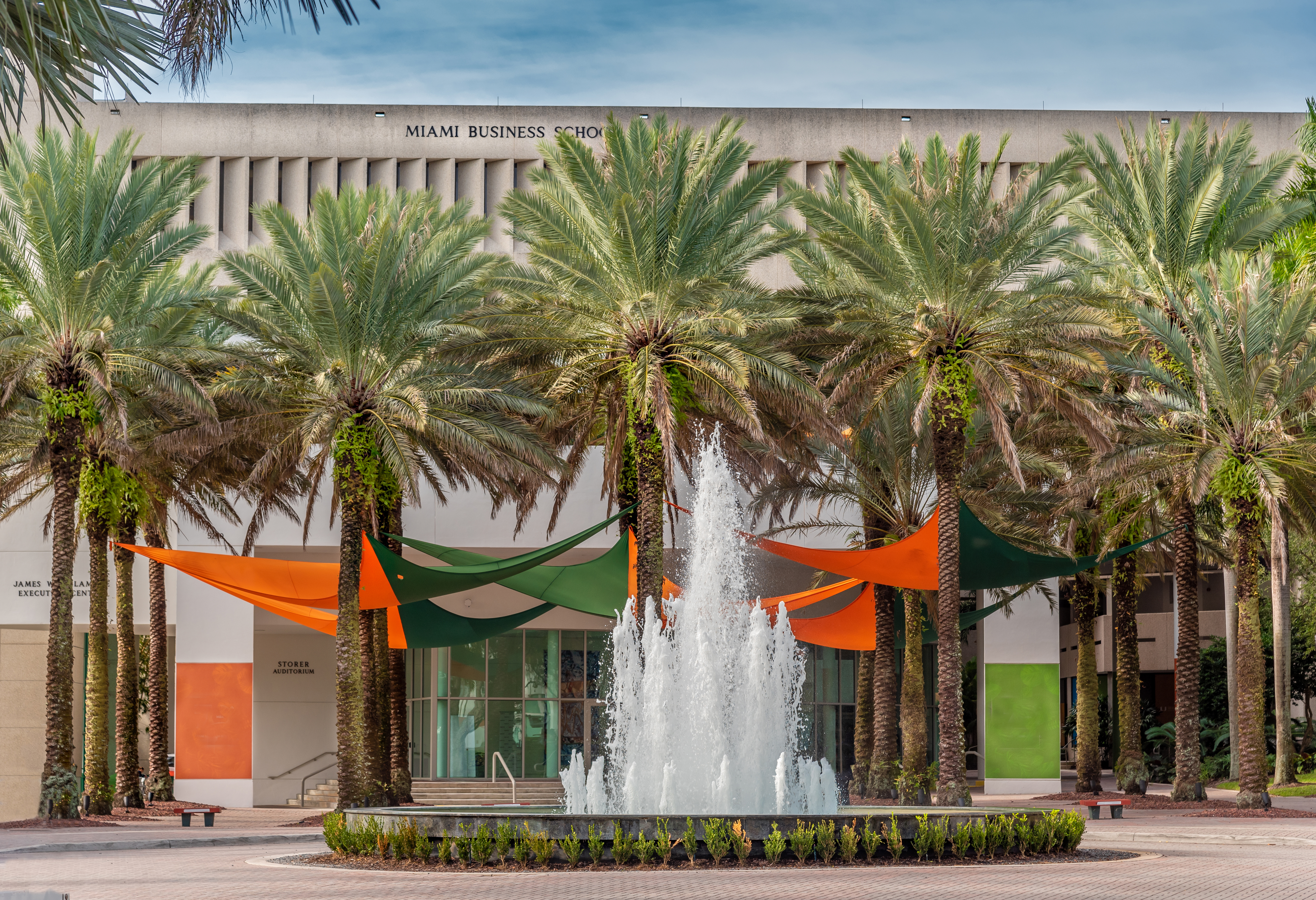 View of a fountain with palm trees, a building, and orange and green banners in the background.
